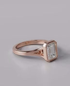 A rose gold ring featuring an emerald-cut clear gemstone, shown on a neutral gray background.