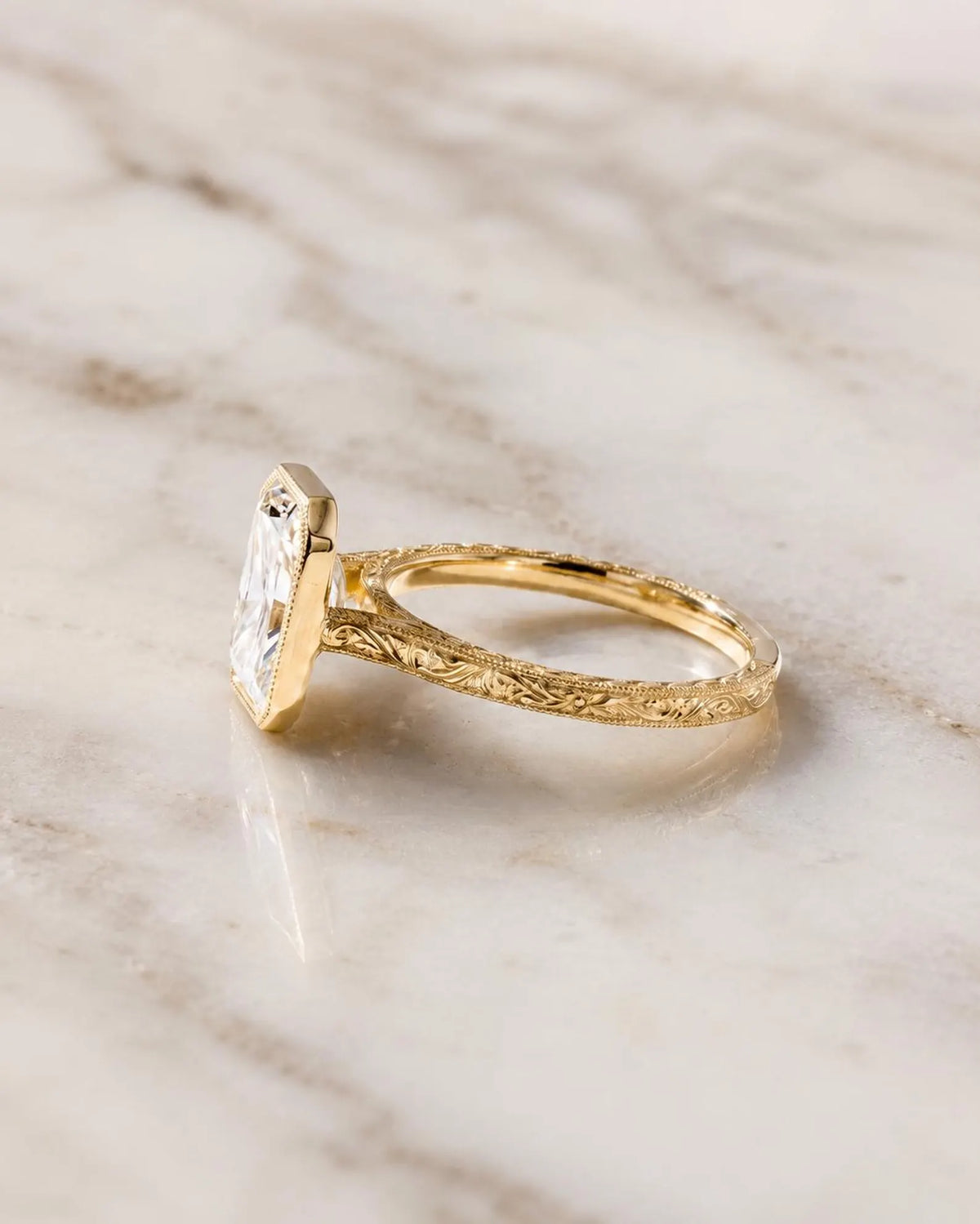 Side perspective view of a 4 CT radiant cut lab grown diamond engagement ring in yellow gold with hand-engraved scrollwork shank.