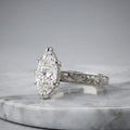Side view of a 4 CT marquise cut lab grown diamond engagement ring with hand engraving in platinum on a marble surface.