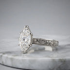 Side view of a 4 CT marquise cut lab grown diamond engagement ring with hand engraving in platinum on a marble surface.