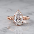 Front view of a 4 CT pear cut lab grown diamond engagement ring with scalloped milgrain halo and engraved rose gold band on marble.