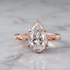 Front view of a 4 CT pear cut lab grown diamond engagement ring with scalloped milgrain halo and engraved rose gold band on marble.