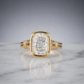 Front view of a 3 CT elongated cushion cut lab grown diamond ring in a yellow gold bezel frame with diamond cuff accents on marble.