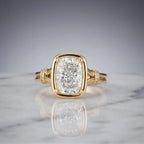 Front view of a 3 CT elongated cushion cut lab grown diamond ring in a yellow gold bezel frame with diamond cuff accents on marble.
