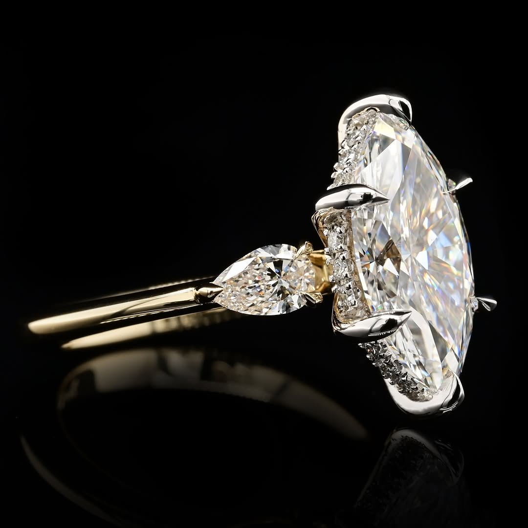 An marquise diamond ring with a yellow gold band, featuring a pavé-set diamond halo and a pear-cut diamond accent on the shank, photographed on a reflective black background.