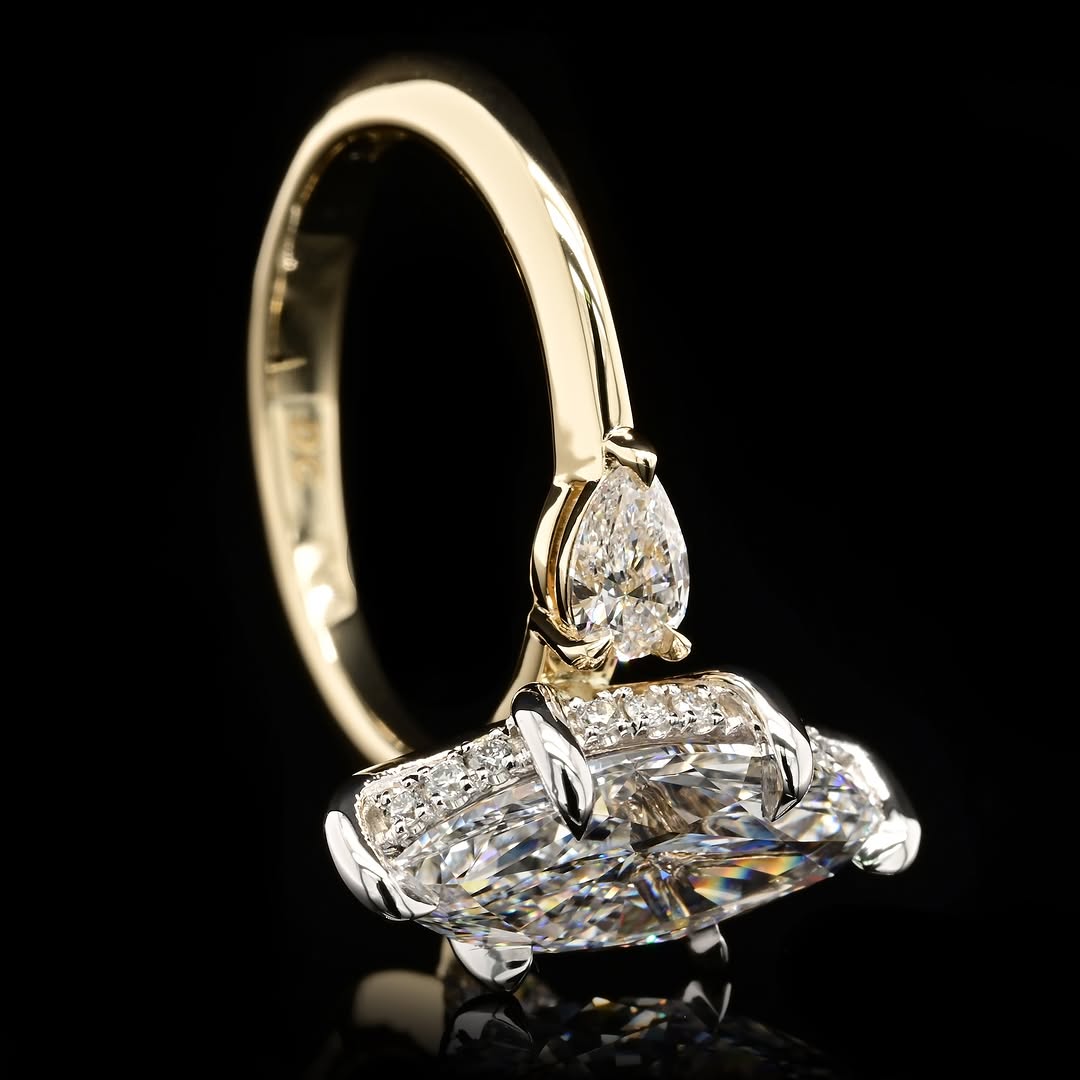 A yellow gold ring featuring a large pear-cut diamond and a larger marquise-cut diamond cluster with pavé accents, set against a solid black background.