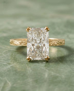Front view of a 4 CT elongated radiant cut lab grown diamond ring with hand-engraved yellow gold band on green marble.
