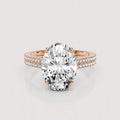 An oval-cut diamond engagement ring in a rose gold setting with a pavé band, shown on a light gray reflective background.