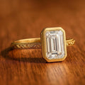 Horizontal view of 3 Carat Emerald Cut Lab Grown Diamond Engagement Ring with hand-engraved band resting on dark wood