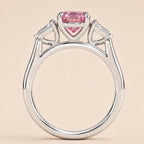A silver-toned ring featuring a heart-shaped pink gemstone at the center, flanked by two small clear stones in triangular settings, shown against a solid pale beige background.