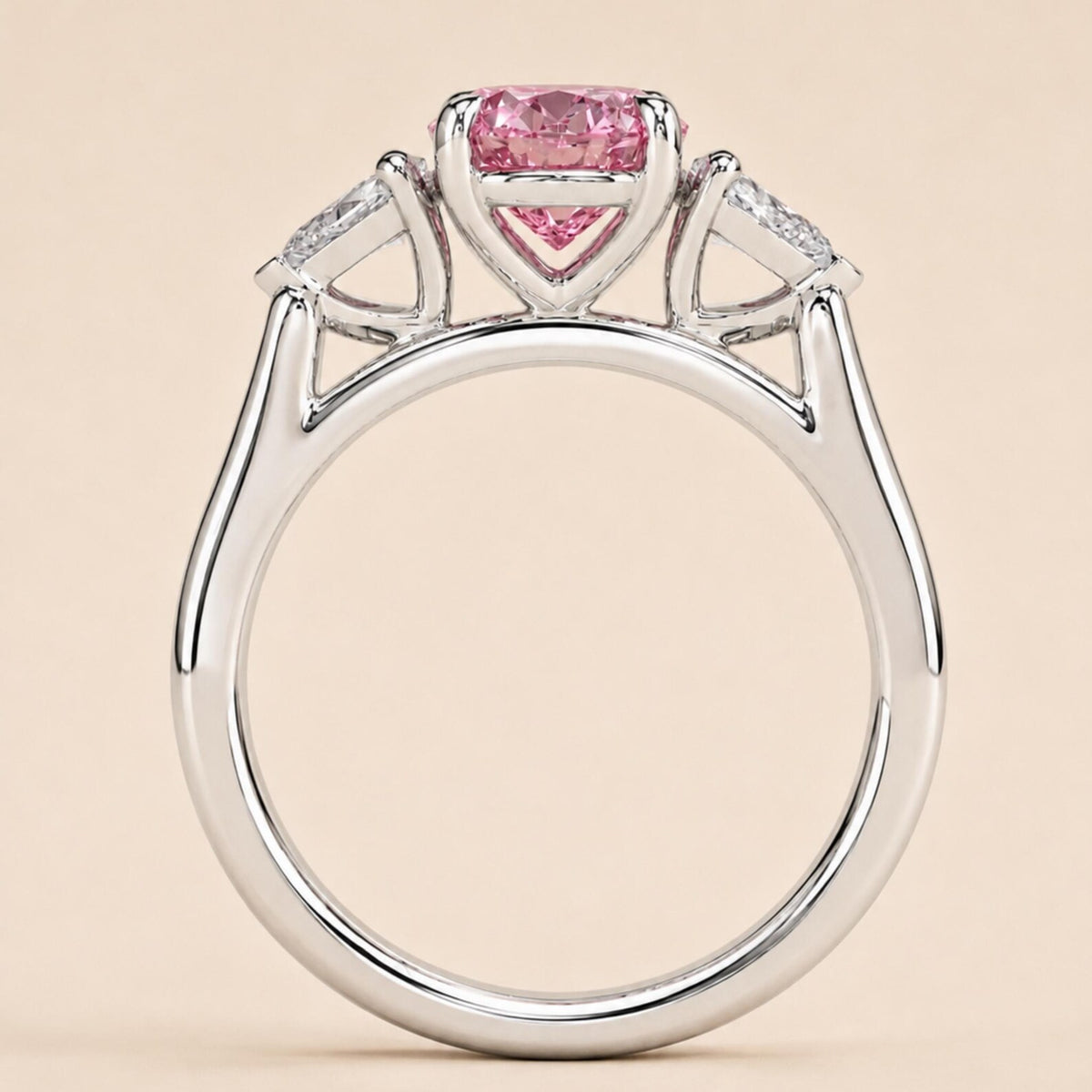 A silver-toned ring featuring a heart-shaped pink gemstone at the center, flanked by two small clear stones in triangular settings, shown against a solid pale beige background.