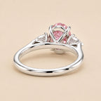 A silver ring featuring a central oval pink gemstone in a prong setting, with small clear accent stones on either side, shown on a warm beige background.