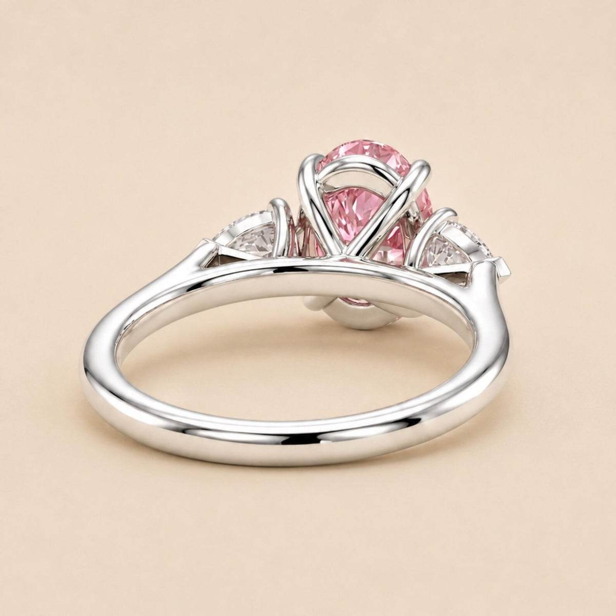 A silver ring featuring a central oval pink gemstone in a prong setting, with small clear accent stones on either side, shown on a warm beige background.