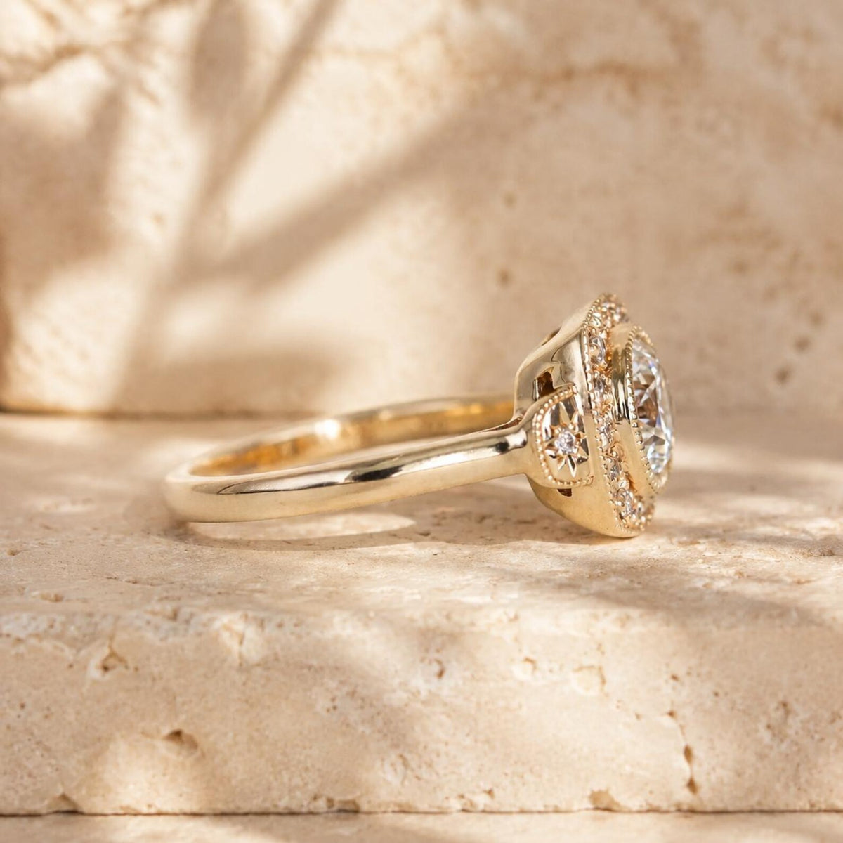 A yellow gold ring with a halo-style setting and a large oval center stone, photographed on a textured beige stone surface with warm sunlight and shadows.