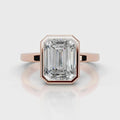 An emerald-cut diamond ring set in rose gold, featuring a prominent rectangular gemstone in an octagonal bezel setting on a polished band, shown against a light gray background with a soft reflection.