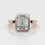 An emerald-cut diamond ring set in rose gold, featuring a prominent rectangular gemstone in an octagonal bezel setting on a polished band, shown against a light gray background with a soft reflection.
