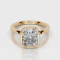 A cushion-cut diamond ring in a yellow gold bezel setting, shown on a clean light gray reflective surface.
