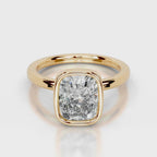 A cushion-cut diamond ring in a yellow gold bezel setting, shown on a clean light gray reflective surface.