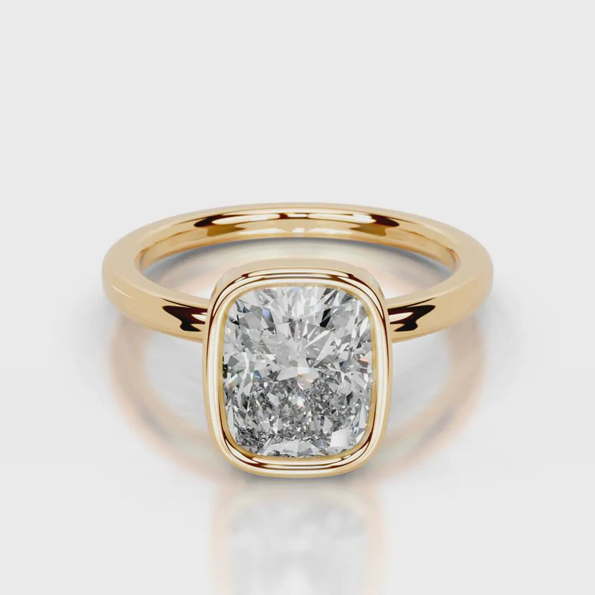A cushion-cut diamond ring in a yellow gold bezel setting, shown on a clean light gray reflective surface.