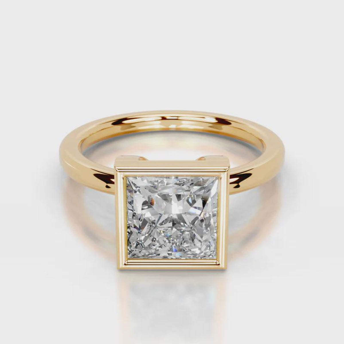 A yellow gold ring featuring a square bezel-set princess-cut diamond, shown on a clean light gray reflective background.
