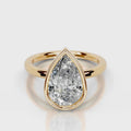 A pear-cut diamond ring in a yellow gold setting with a split shank band, shown on a light gray reflective surface.