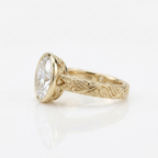 Side profile angle of a yellow gold 3 ct oval lab diamond bezel ring showing hand-carved leaf shank details.