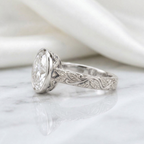 Side angle view of a white gold bezel set 3 ct oval lab grown diamond ring with carved leaf pattern shank.