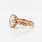 Side profile angle of a rose gold 3 carat oval lab diamond bezel ring featuring intricate leaf hand engraving.