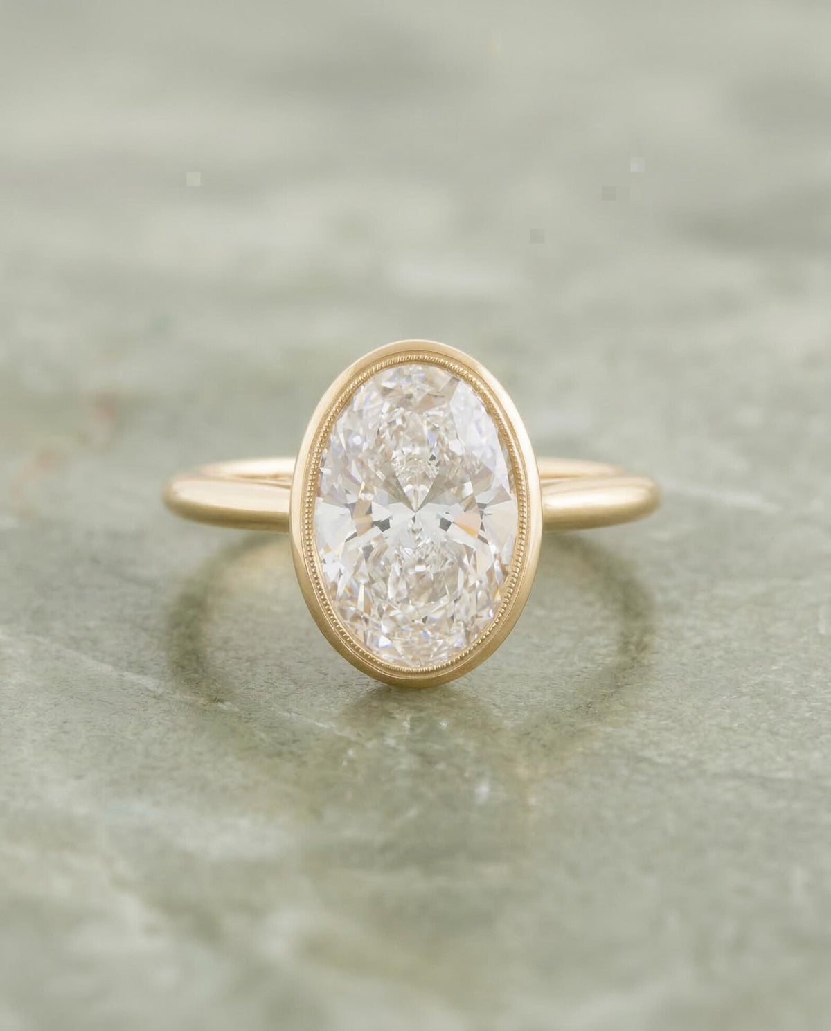    Top-down view of a 2 CT oval cut lab grown diamond engagement ring in a yellow gold bezel setting with milgrain trim.