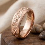 Macro detail shot of the handcrafted floral scroll motif on a rose gold men's wedding ring resting on wood.