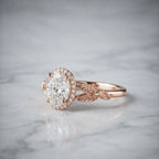 Side profile angle of a rose gold 2 CT oval lab diamond halo ring featuring leaf engraved split shoulders on marble.
