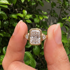 Close-up view held between fingers showing a 5 CT radiant cut lab grown diamond ring with bezel side stones.