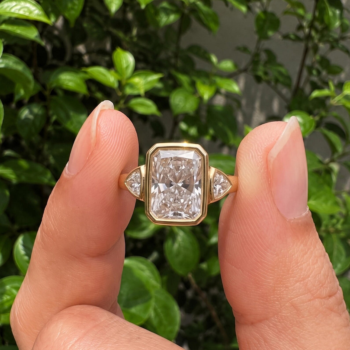 Close-up view held between fingers showing a 5 CT radiant cut lab grown diamond ring with bezel side stones.