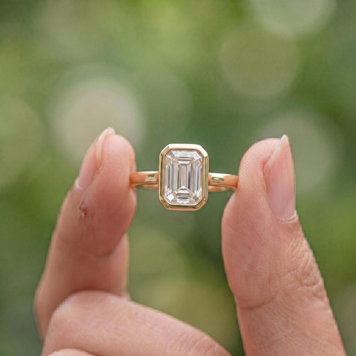 Close-up view of a 2 CT emerald cut lab diamond bezel ring held between fingers outdoors.