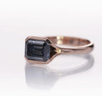Side angle view of a 2 CT grey emerald cut moissanite engagement ring in a rose gold bezel setting.