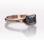Angled view showcasing the grey emerald-cut moissanite center stone and smooth rose gold band.