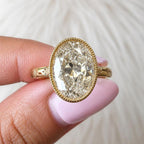 On-hand top view of a 5 carat oval cut lab diamond encased in a detailed yellow gold milgrain bezel setting.