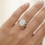 A close-up of a hand wearing a yellow gold ring set with a oval-cut diamond and small accent diamonds on a matching band, against a soft light beige background.