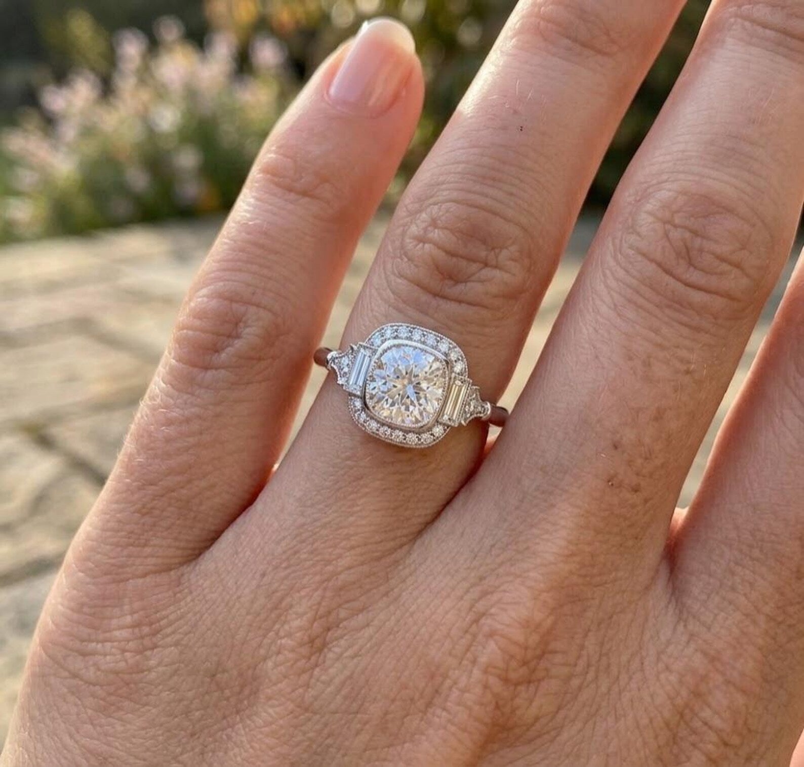 Woman's hand wearing a stunning vintage-style 3 CT cushion cut lab grown diamond engagement ring with baguette side stones in natural outdoor lighting.