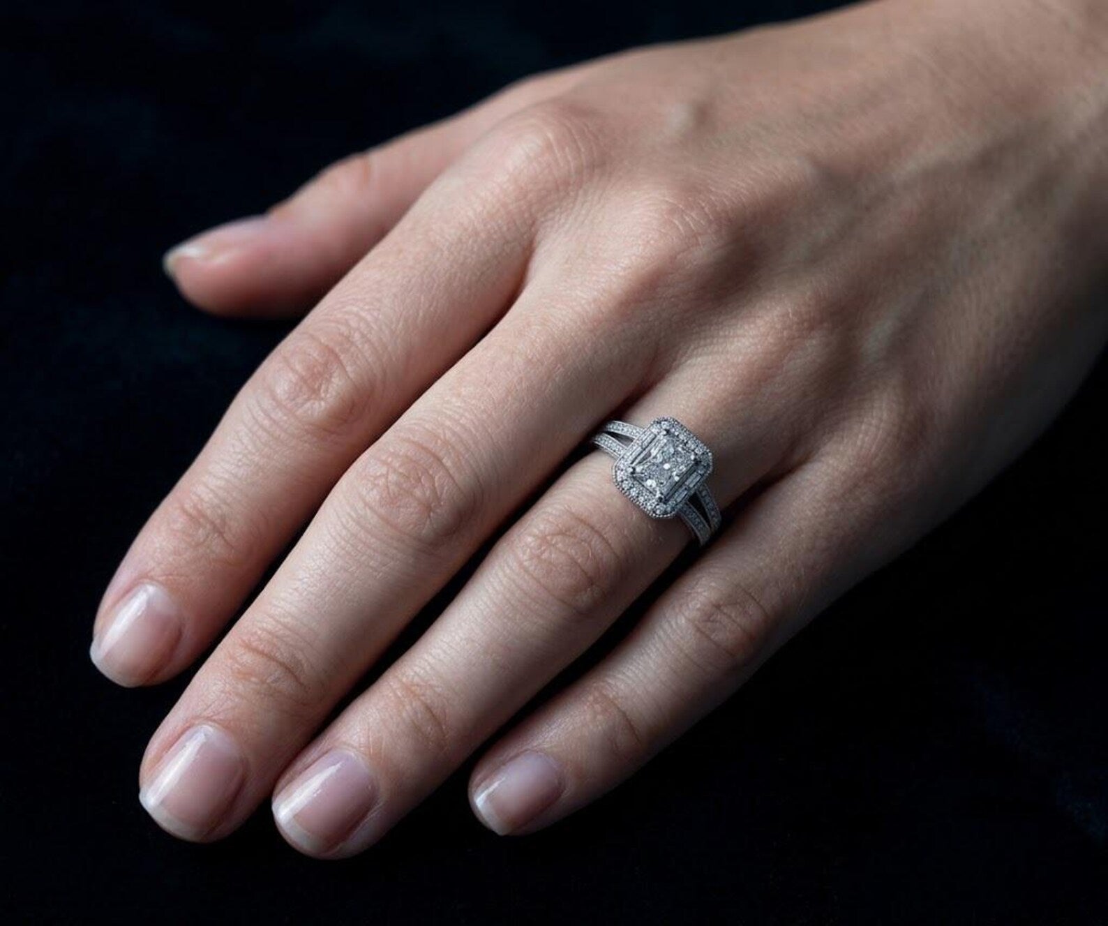 Lifestyle image of a woman's hand wearing a spectacular 3 CT radiant cut split shank halo engagement ring against a contrasting dark background.
